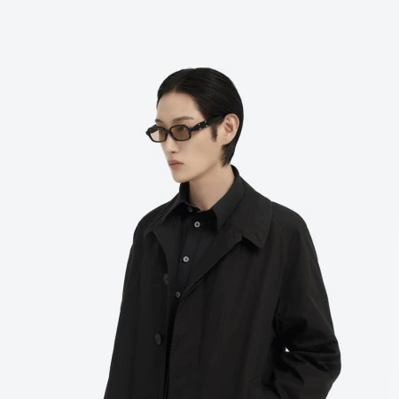Close-up studio shot of an adult with dark hair wearing a black shirt and dark coat with a pair of retro framed glasses.