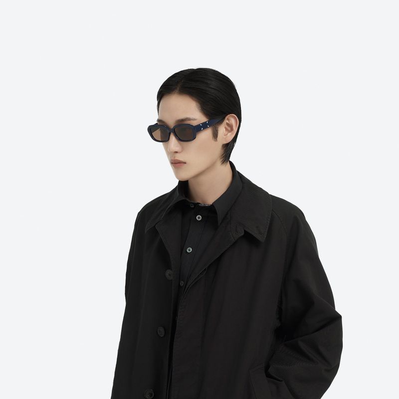 A stylish person with dark hair wearing black collared shirt, black coat and designer sunglasses against a plain white backdrop.