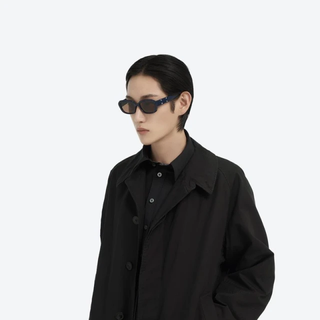 A stylish person with dark hair wearing black collared shirt, black coat and designer sunglasses against a plain white backdrop.