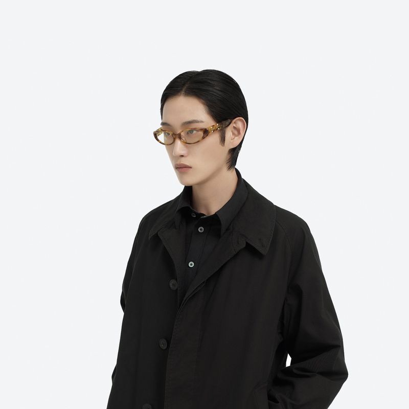 A person wearing brown-framed glasses and a black coat poses in a minimalist white studio environment, highlighting fashion and style.