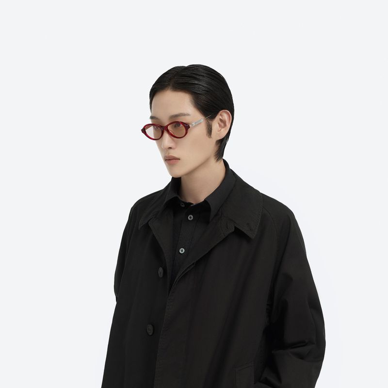 Portrait of a young adult in a black shirt and coat, wearing oval red eyeglasses in a brightly lit photo studio environment.