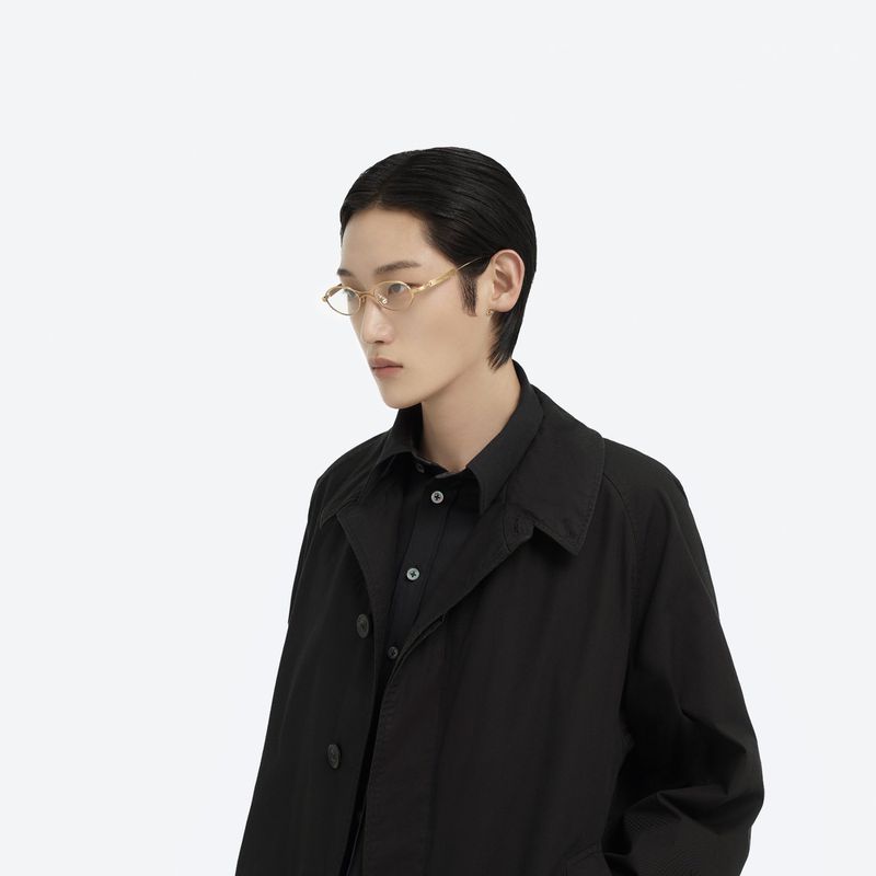 Model wearing gold wire-frame glasses and black coat against white background for Maison Margiela X Gentle Monster