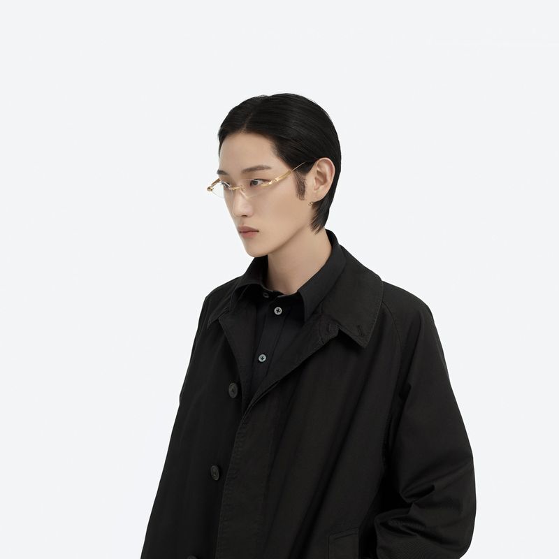 Man in a black collared shirt and dark coat poses in front of a white backdrop, wearing thin-framed gold glasses.