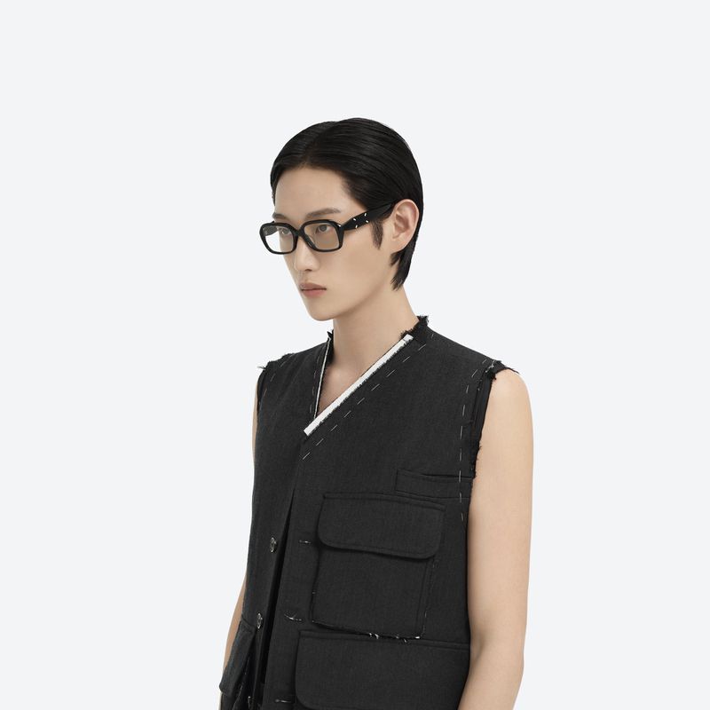 Headshot of an adult with short, dark hair wearing black framed glasses and a dark sleeveless vest against a white backdrop.