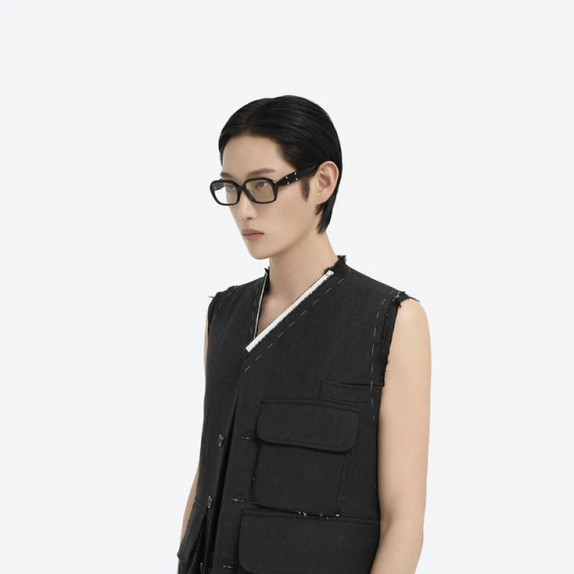 Headshot of an adult with short, dark hair wearing black framed glasses and a dark sleeveless vest against a white backdrop.