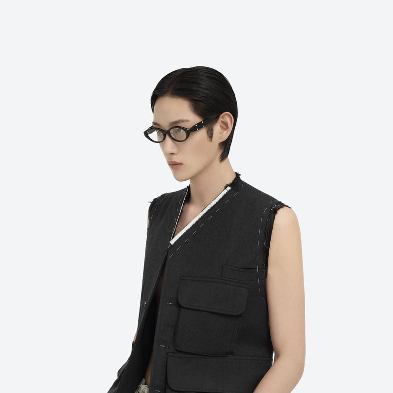Person with short dark hair wears black-framed glasses and a black utility vest against a bright white studio backdrop in a medium shot.