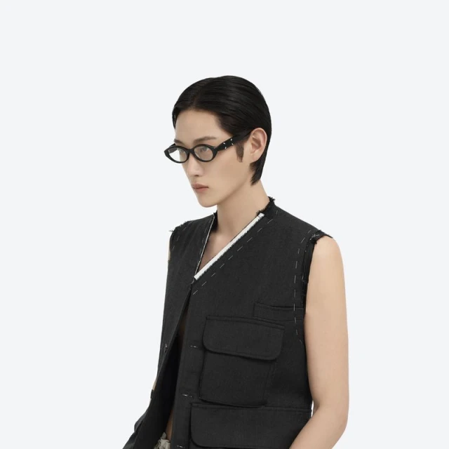 Person with short dark hair wears black-framed glasses and a black utility vest against a bright white studio backdrop in a medium shot.