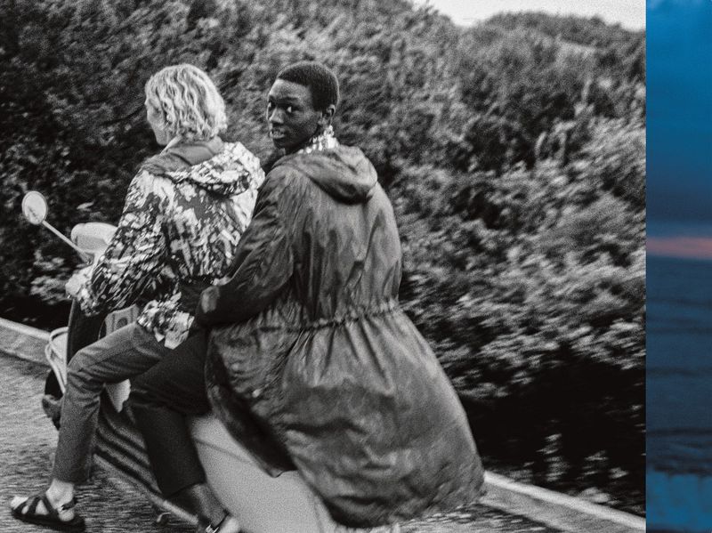 Two people riding a scooter on a road. The Black male passenger is looking at the camera, wearing a parka. Trees are visible in the background.
