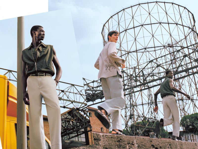 Three people in stylish clothing pose in an urban setting near industrial structure under a blue sky, conveying a modern, fashionable aesthetic.