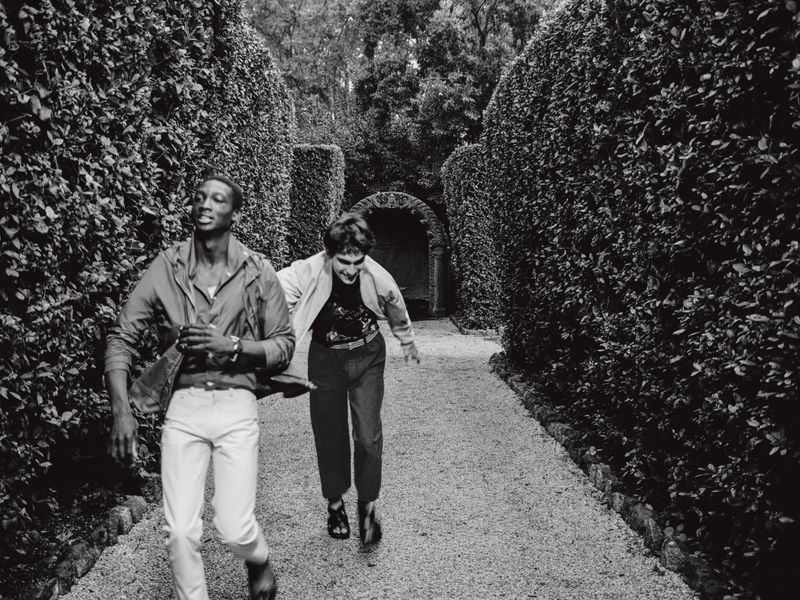 Two people run along a gravel path lined with manicured hedges in a black and white image depicting motion and an outdoor setting.