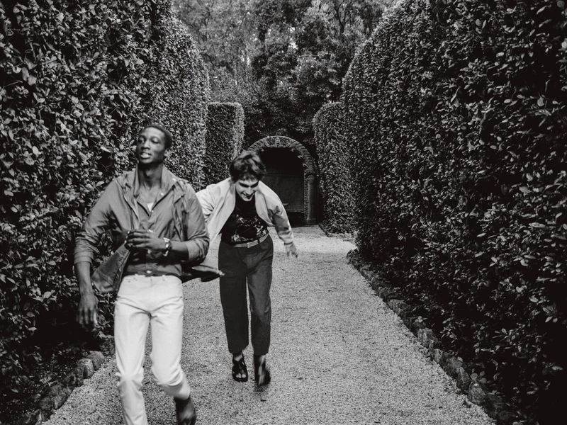 Two people run along a gravel path lined with manicured hedges in a black and white image depicting motion and an outdoor setting.