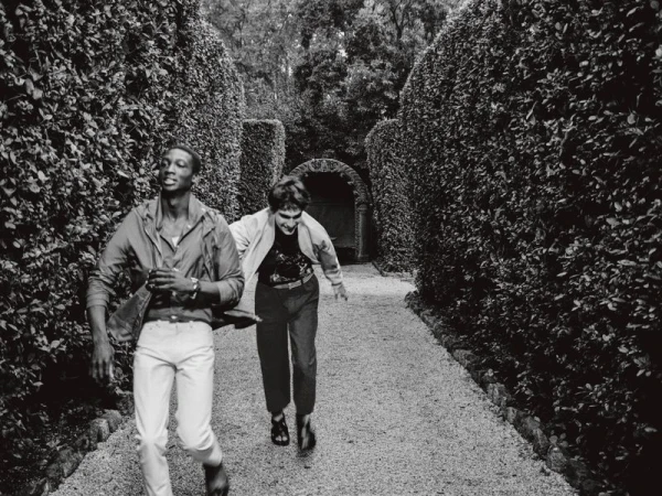 Two people run along a gravel path lined with manicured hedges in a black and white image depicting motion and an outdoor setting.