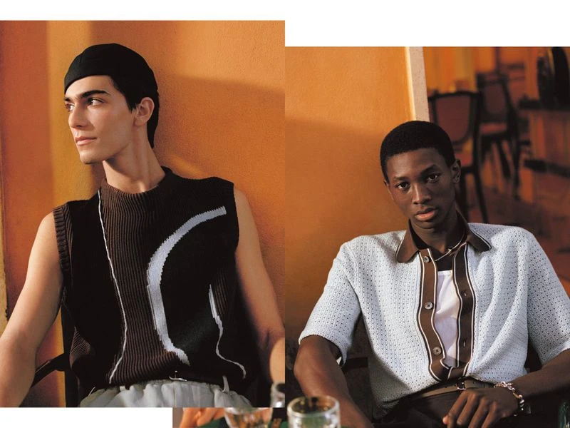 Two male fashion models in stylish knitwear, photographed indoors with an orange wall backdrop in a contemporary editorial style.
