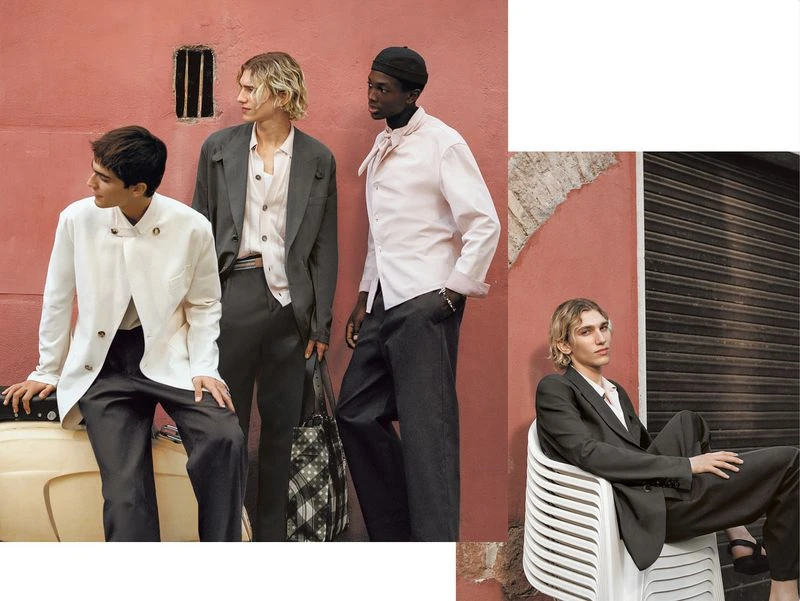 Three fashion models in suits pose against a coral-colored wall with a vintage moped. A fourth model in a suit sits in a white chair.