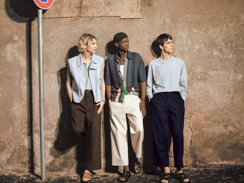 Three stylish men in casual summer outfits pose against a textured wall with a traffic sign, showcasing urban fashion trends.
