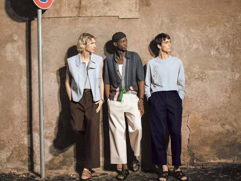 Three stylish men in casual summer outfits pose against a textured wall with a traffic sign, showcasing urban fashion trends.