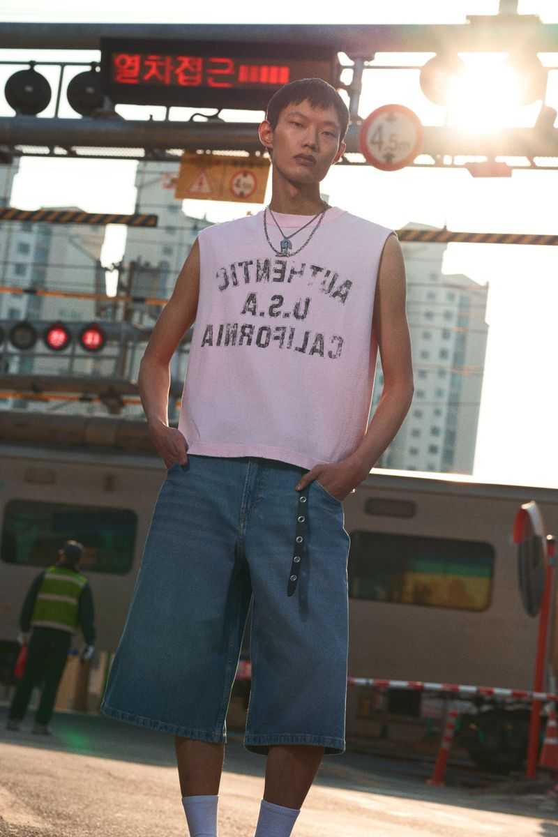 Asian male model in casual fashion stands near railroad tracks with train, wearing pink tank top, blue culottes in outdoor setting.
