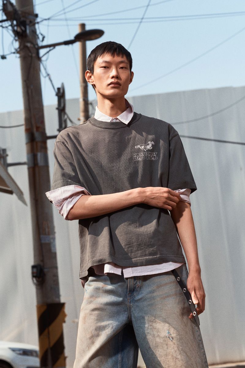 Asian man in gray graphic T-shirt layered over a light pink collared shirt, with distressed blue jeans, posing in an urban setting outdoors.