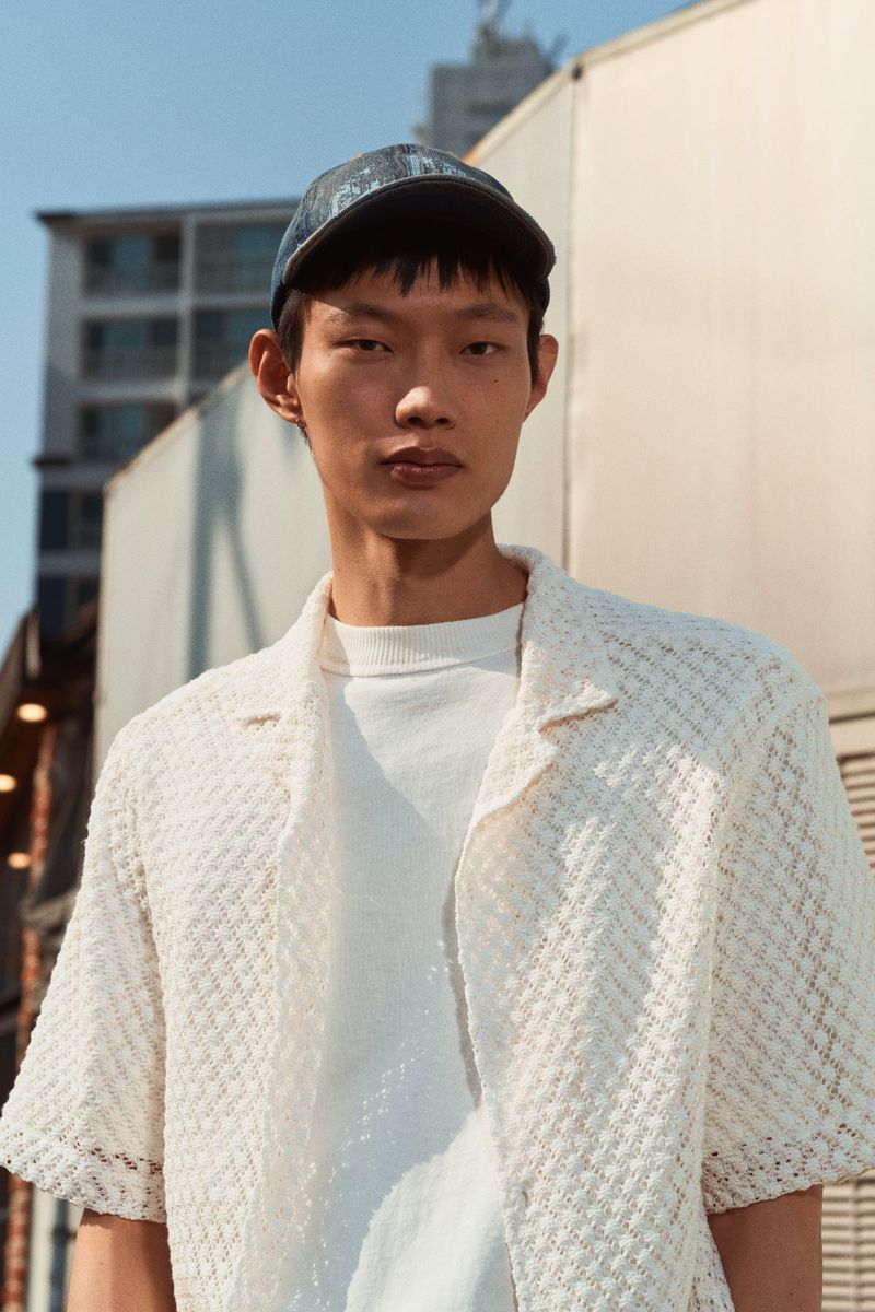 Asian man wearing a white shirt and baseball cap standing outside on a sunny day with urban buildings in the background.