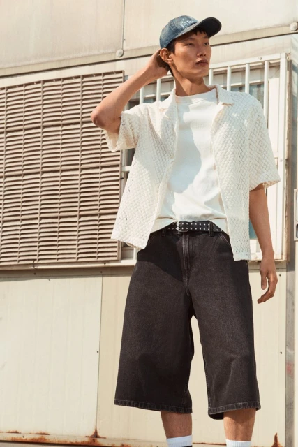 Man in streetwear fashion posing outdoors. He wears a baseball cap, open white textured shirt, white t-shirt, dark denim shorts, and sneakers.