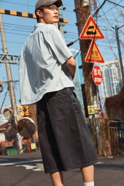 Man wearing a light blue striped shirt, dark denim shorts, a baseball cap, and socks, stands on a city street with signs and buildings.