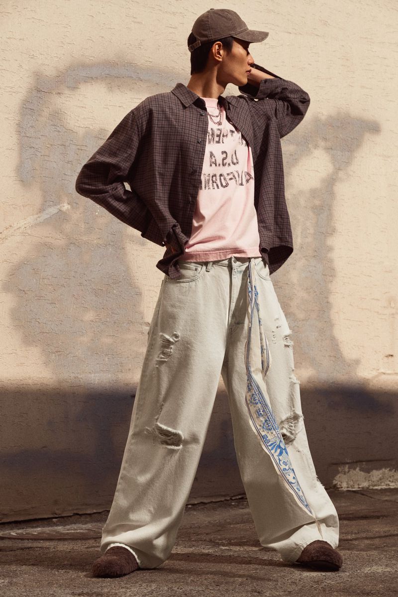 A young adult man in casual streetwear poses in front of a wall, wearing a cap, unbuttoned shirt, t-shirt, baggy jeans, and moccasin-style shoes.