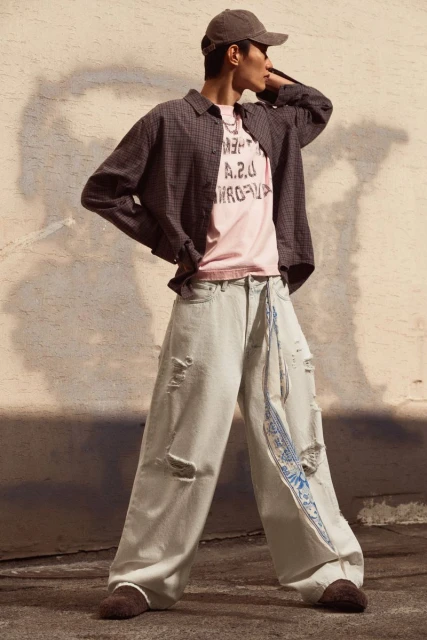 A young adult man in casual streetwear poses in front of a wall, wearing a cap, unbuttoned shirt, t-shirt, baggy jeans, and moccasin-style shoes.