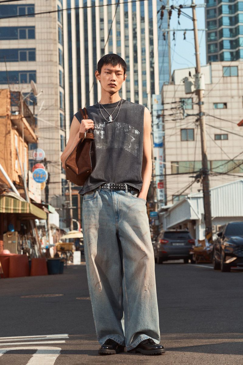 Eye-level shot of a young adult male in sleeveless shirt and baggy jeans standing on a city street, holding a brown bag on his shoulder.