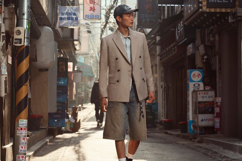 Stylish adult male strolls down a narrow urban street, wearing a beige blazer, blue shirt, denim shorts, and a baseball cap in daytime.