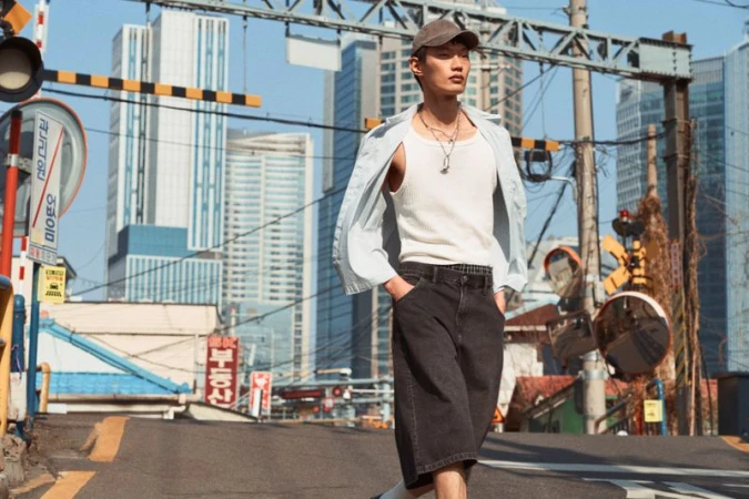 Asian man in casual streetwear stands on city street with modern skyscrapers and old buildings in background on a sunny day.