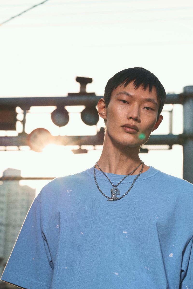 An Asian male model is posing outdoors wearing a blue t-shirt and necklaces with an urban backdrop and soft sunlight.
