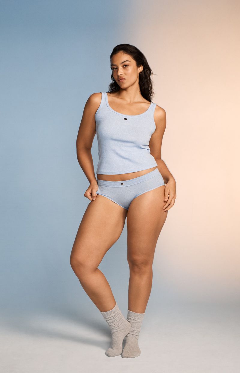 Full shot of a woman in a light blue tank top and underwear, standing in front of a gradient blue and peach background in a studio.