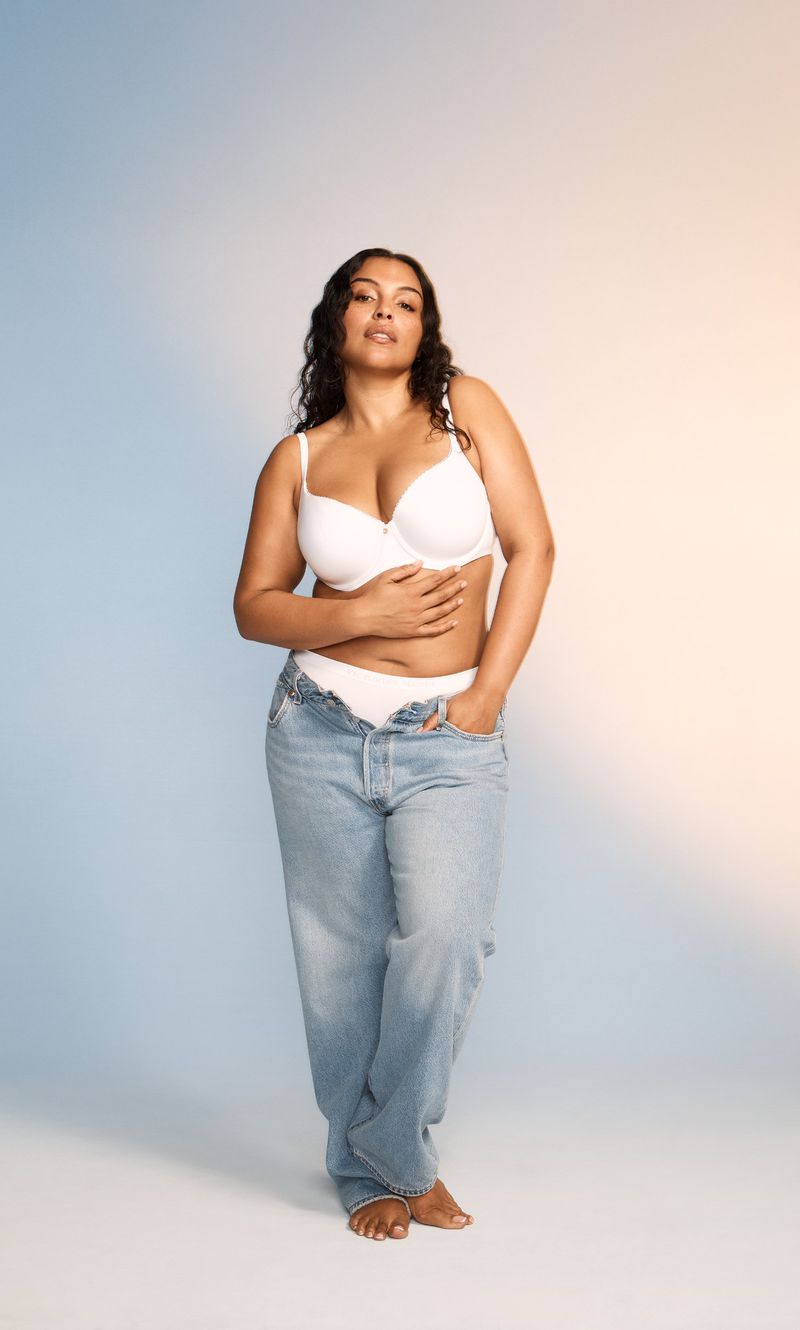 Portrait of an adult woman with curly hair posing in a bra and light-wash jeans against a gradient blue and beige background.