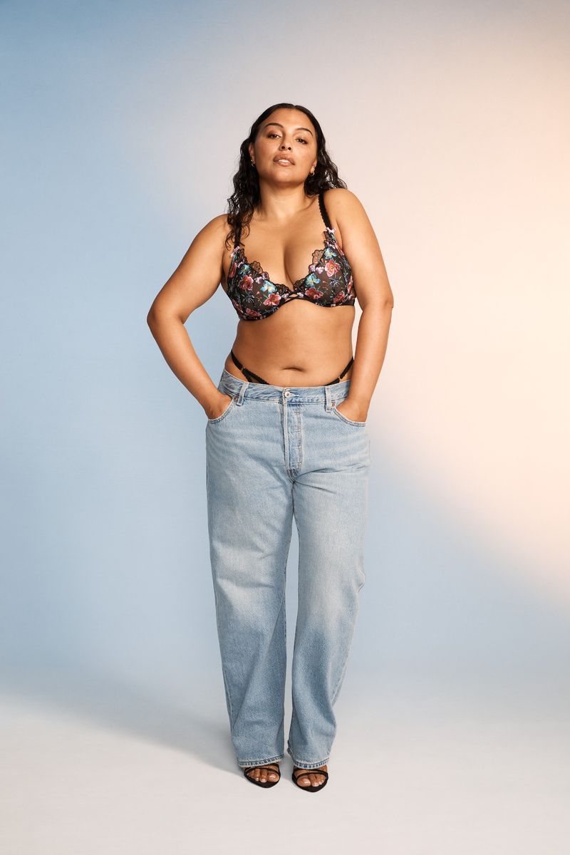 Full-length portrait of a female model posing in a floral bra top and jeans, hands in pockets, in a photo studio with gradient lighting.