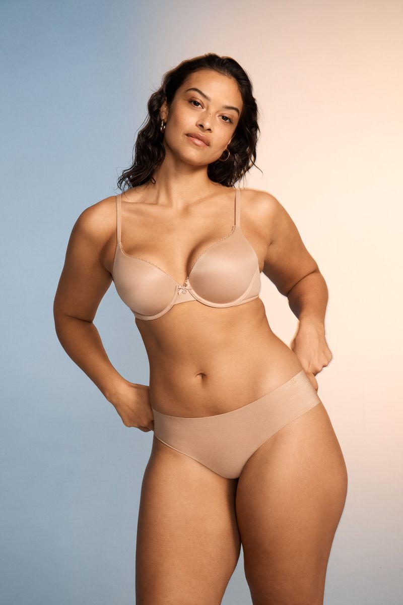 Woman in bra and underwear poses against a blue and beige gradient background in a medium shot. Lingerie product photography.