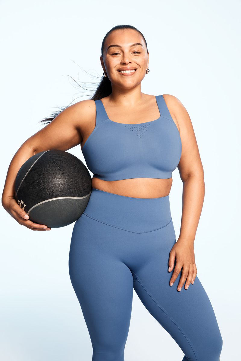 Athletic woman in sports bra and leggings holds medicine ball against a white background, smiling and looking at the camera.