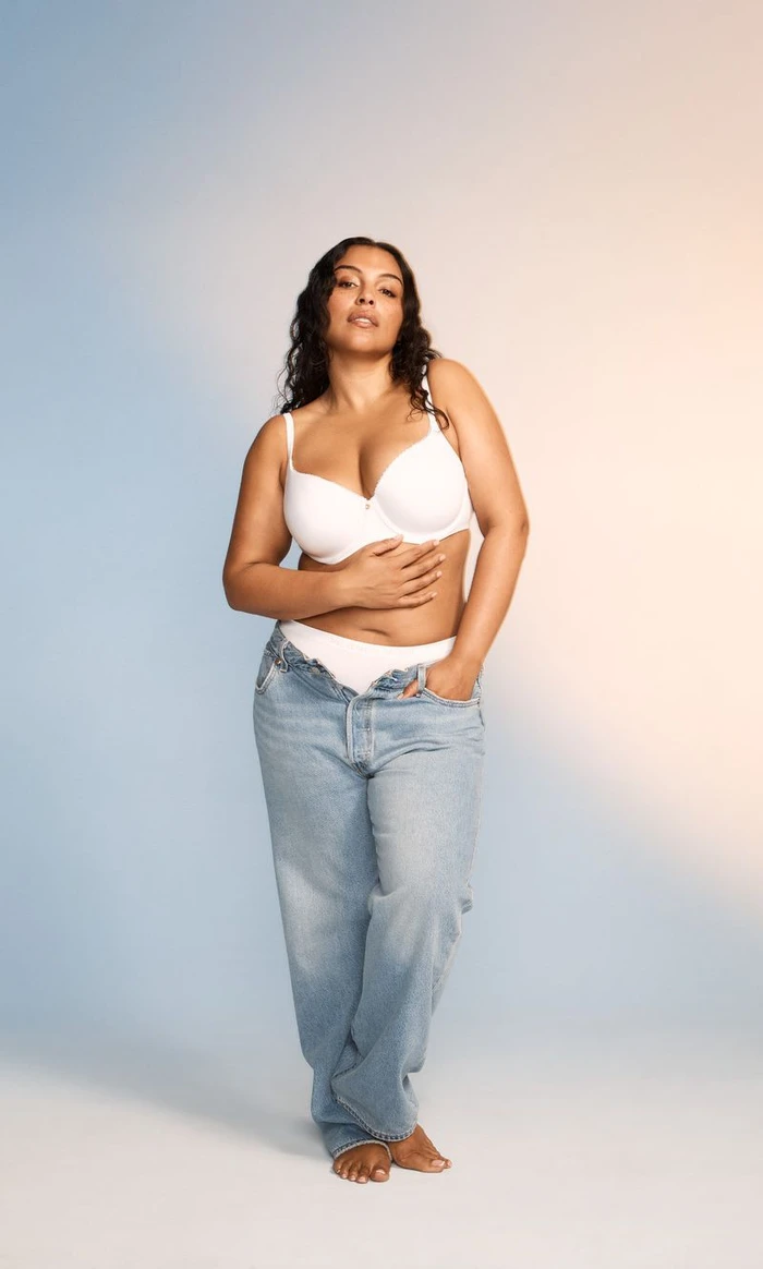 Model wearing white bra and unbuttoned denim jeans against a soft blue and warm gradient backdrop