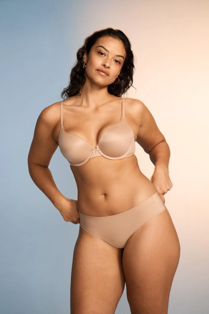 Model wearing nude lingerie standing against a gradient blue and warm light studio background.