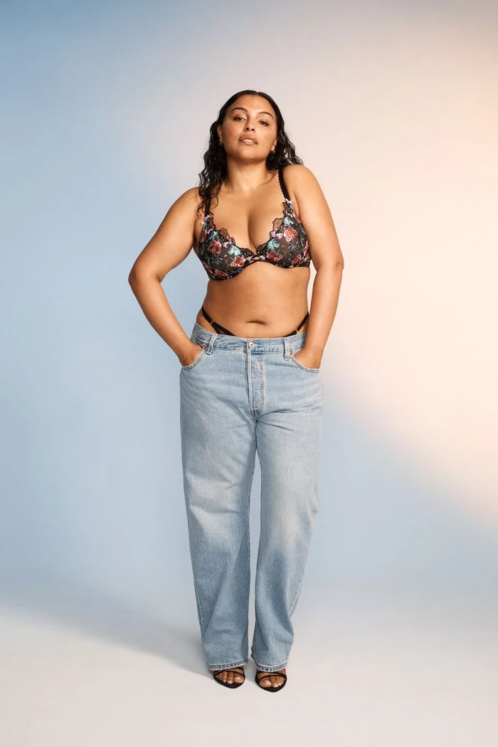 Model wearing a floral lace bra and light wash denim jeans standing against a blue to orange gradient backdrop.