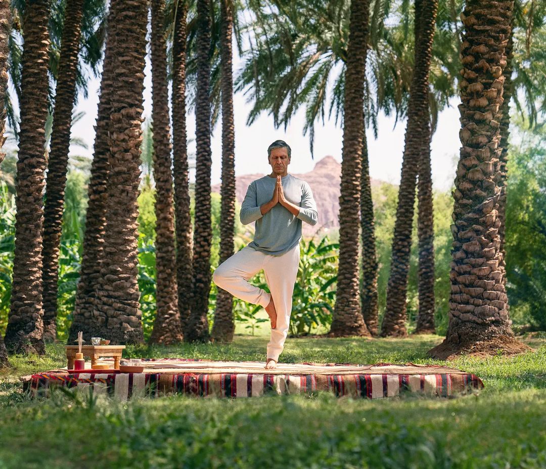 Adult male practices yoga pose on a red patterned rug in a tranquil outdoor setting surrounded by tall palm trees and greenery.