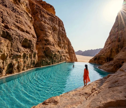Woman in red dress standing beside a long outdoor pool in a rocky desert canyon; blue water contrasts against the tan rocks and sand.