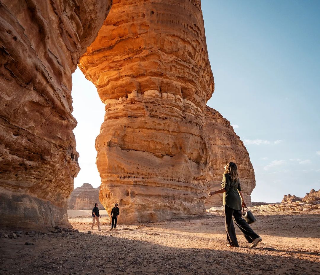 Woman walks through desert landscape with dramatic sandstone rock formations and two figures in the distance under a clear blue sky.
