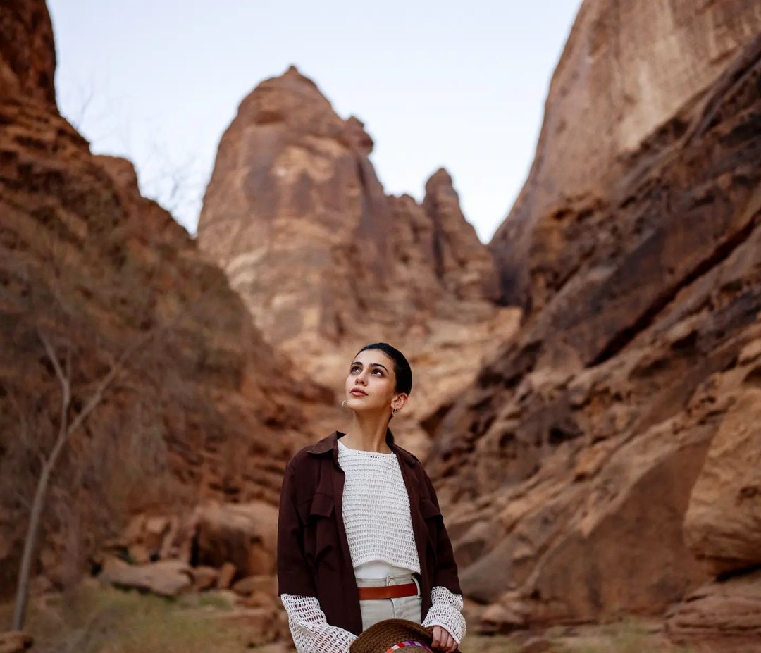 Woman in brown jacket looks up in desert canyon, lit by soft light, wearing white sweater and holding hat. Travel and fashion photography.