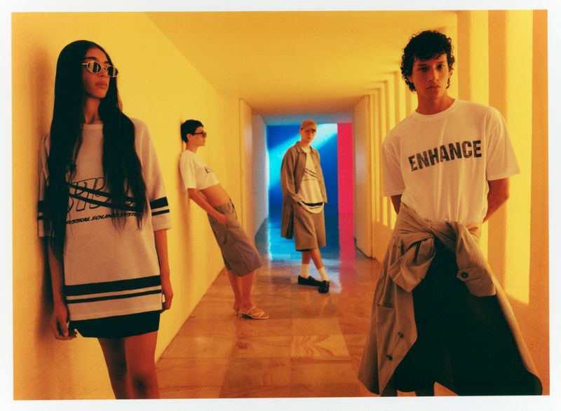 Four fashion models showcase contemporary apparel in a bright hallway, featuring t-shirts, shorts, and stylish accessories in a studio setting.