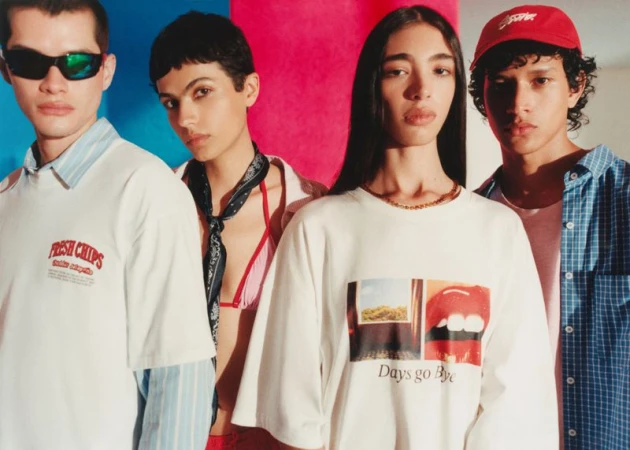 Four young adults pose indoors against a vibrant pink and blue wall. Two wear white graphic tees, one a red cap, and another sunglasses.
