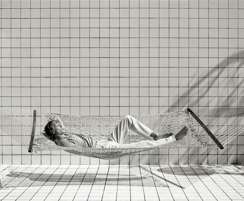 Adult lies in a net hammock indoors on tiled floor and walls, dappled by shadow and light in a black and white lifestyle photograph.