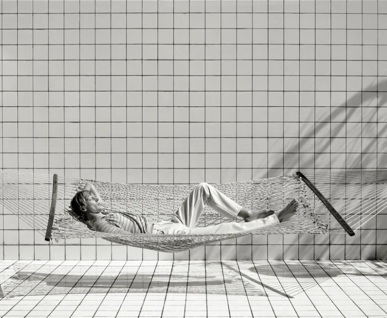 Adult lies in a net hammock indoors on tiled floor and walls, dappled by shadow and light in a black and white lifestyle photograph.