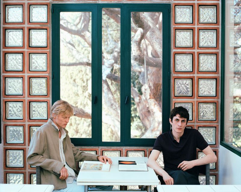 Two men seated at a table indoors, looking at books or documents, with a window and textured glass block wall behind them.