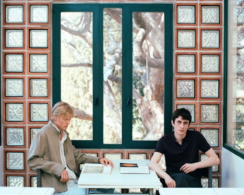 Two men seated at a table indoors, looking at books or documents, with a window and textured glass block wall behind them.