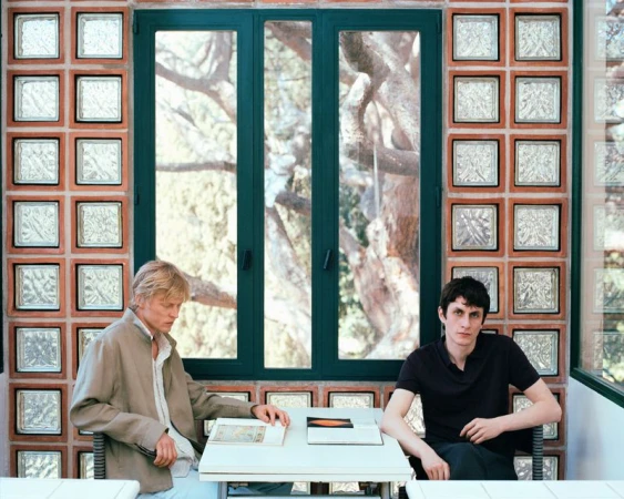 Two men seated at a table indoors, looking at books or documents, with a window and textured glass block wall behind them.